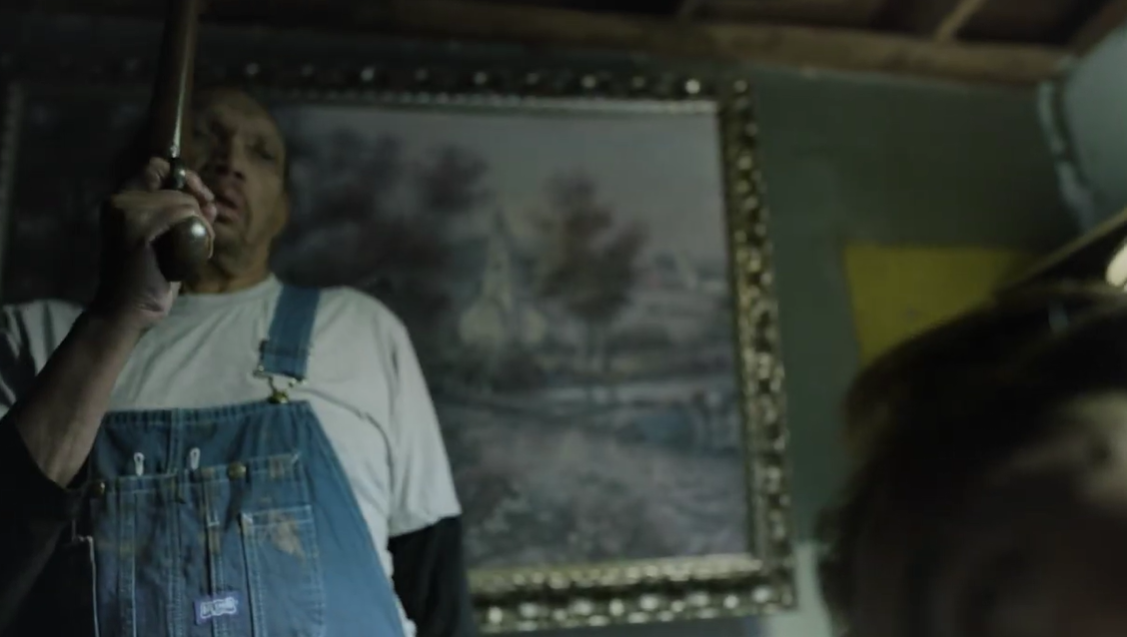 Film still: a man in overalls beneath a framed landscape painting in a dim farmhouse